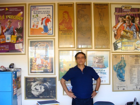 Luiz Carlos Ribeiro em seu escritório na sede da Cia Teatro de Risco, com os quadros dos espetáculos, ao fundo